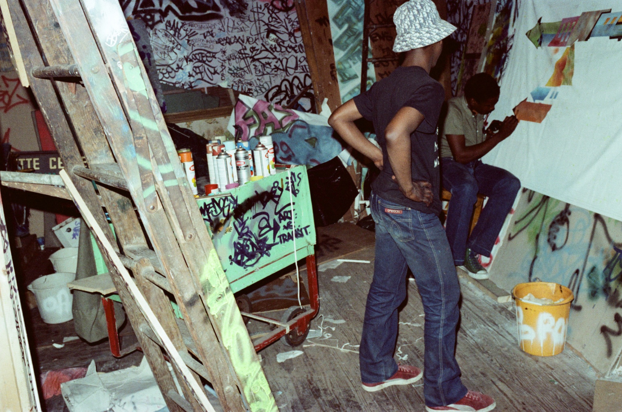 A-One watching Kool Koor creating his first artwork on canvas at Fashion Moda, 1982